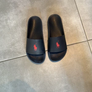 Svarta slides från Polo Ralph Lauren strl 41,5 - Svarta sandaler från Polo Ralph Lauren med röd logotyp framtill. De har en bred rem över foten och en skön, strukturerad sula i syntetmaterial. Perfekta för sommaren eller att chilla i hemma.