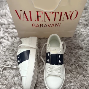 Valentino Garavani vita sneakers - Snygga vita Valentino Garavani sneakers med bred blå rem över vristen och coola nitar på sidorna. Skorna har rund tå, sula med mönstrad undersida. De är i fint skick men boxen finns inte kvar (den var sliten), därför har jag sänkt priset rejält.