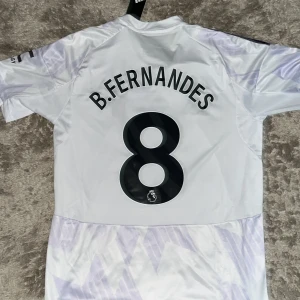 Manchester United B. Fernandes tröja S - Säljer en officiell Manchester United bortatröja från Adidas med B. Fernandes och nummer 8 på ryggen. Tröjan är vit med ljuslila mönster, svarta detaljer och klassiska Adidas-ränder på axlarna. Materialet är lätt och andas, perfekt för match eller träning.