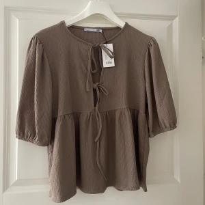 Brun blus med knyt och puffärm - Superfin brun blus med puffiga trekvartsärmar och knyt framtill. Blusen har en lätt rynkad, flowy passform och är gjord i ett mjukt, strukturerat tyg. Perfekt för dig som gillar en avslappnad men ändå söt stil.