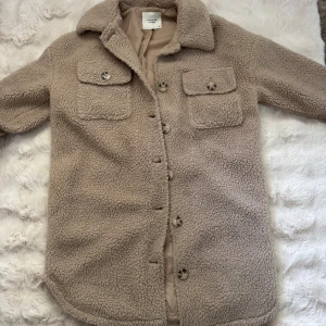 Beige teddyjacka från Jacqueline de Young - Mysig beige teddyjacka från Jacqueline med två stora bröstfickor och knäppning framtill. Jackan har klassisk krage, lång ärm och är tillverkad i fluffigt teddy-material som håller dig varm och stylish. Perfekt för lager-på-lager och chill höst- och vinterdagar.