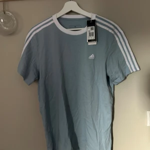 Blå Adidas t-shirt med vita ränder - Snygg ljusblå t-shirt från Adidas med klassiska vita ränder längs axlarna och ärmarna. T-shirten har rund hals med vit kant och Adidas-logga på bröstet. Perfekt för dig som gillar sportig stil och vill ha något bekvämt i bomull.