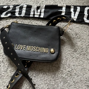 Svart axelväska från Love Moschino - Cool svart axelväska från Love Moschino i skinn med guldiga detaljer och nitar på axelremmen. Väskan har ett stilrent uttryck med logga i guld framtill och bred axelrem. Perfekt för dig som vill ha en edgy och trendig accessoar. Tillkommer även ett band där det står ”Love moschino”