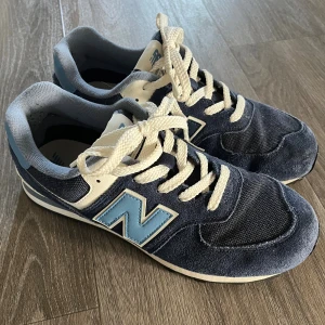 Blå New Balance sneakers strl 39 - Snygga New Balance sneakers i mörkblått med ljusblå och vita detaljer. Skorna har klassisk snörning, rund tå och platt sula. Ovandelen är i mocka och textil, med den ikoniska N-loggan på sidan. 