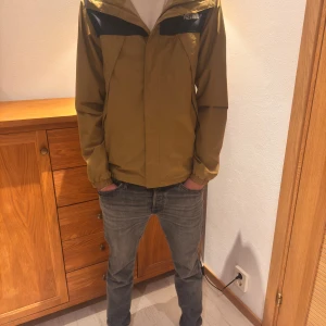Olivgrön vindjacka från The North Face - Säljer en olivgrön vindjacka från The North Face med svarta detaljer över axlarna och huva. Väl använd men inga defekter! Superbra som all day jacka men den är också regntät och vindtät!💗