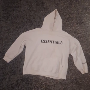 Vit Essentials hoodie med huva - Vit hoodie från Essentials Fear of God med svart tryck på bröstet och ärmen. Klassisk känguruficka framtill, ribbade muddar och stor huva. Perfekt för en clean och avslappnad streetstyle-look.