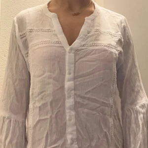 Vit långärmad skjorta med spetsdetaljer - Säljer en vit långärmad skjorta med snygga spetsdetaljer och knappar framtill. Skjortan har en v-ringad hals och är tillverkad i ett lätt, luftigt material som passar perfekt för varmare dagar. Stilren och enkel att matcha med dina favoritjeans eller kjol.