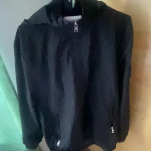 Svart vindjacka från Moncler med huva - Svart vindjacka från Moncler med klassisk logga på ärmen. Jackan har huva, dragkedja framtill och diskreta fickor med dragkedja. Snygg och stilren modell i lätt polyester, perfekt för blåsiga dagar. Passformen är normal och jackan är helsvart utan mönster.