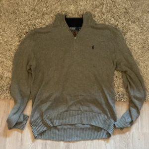 Grå stickad tröja Polo Ralph Lauren - Snygg grå stickad tröja från Polo Ralph Lauren med halv dragkedja och ribbade muddar. Klassisk broderad logga på bröstet och hög krage. Perfekt för lager-på-lager under kyliga dagar.