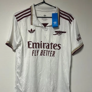 Vit Arsenal fotbollströja Adidas S - Snygg vit Arsenal fotbollströja från Adidas med vinröda detaljer och klassisk krage. Tröjan har korta ärmar, broderade loggor och tryckta sponsor- och klubbmärken. , perfekt för match eller träning.