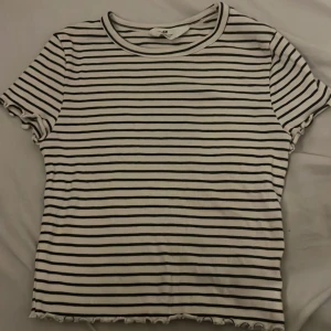 Randig ribbad topp från H&M - Säljer en vit och svart ribbad topp från H&M med smala horisontella ränder. Toppen har korta ärmar, rund halsringning och vågiga kanter vid ärmslut och nederkant. Perfekt för en chill och stilren look. 