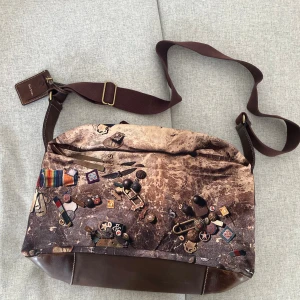 Paul Smith axelväska med print - Unik axelväska från Paul Smith med coolt tryck av små föremål på framsidan. Väskan har bruna skinn- och textildetaljer, justerbar axelrem och innerfoder i lila. Botten och detaljer är i brunt skinn, med dragkedja och flera fack inuti.