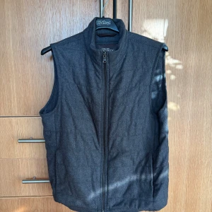 Mörkgrå dunväst från Oscar Jacobson - Snygg mörkgrå dunväst från Oscar Jacobson med hög krage och dragkedja framtill. Västen har en quiltad look, två sidfickor och två innerficka med dragkedja. Utsidan är i ullblandning och fodret är slätt med logga. Perfekt för lager-på-lager under kyliga dagar. Storleken är 48 men kan tolkas som M. Pris är inte fast så bara att skriva om du har ett annat förslag!