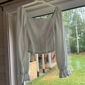 Vit smockad blus med volangärm - Supersöt vit blus med smockad kropp och långa, luftiga ärmar som avslutas med volang. Toppen har en fyrkantig ringning och är croppad, perfekt att styla med höga jeans eller kjol. Materialet är lätt.