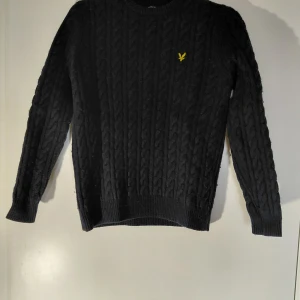 Svart kabelstickad tröja Lyle & Scott - Svart kabelstickad tröja från Lyle & Scott med gul logga på bröstet. Rund hals och ribbade muddar vid ärmslut och nederkant. Mjuk och varm, passar till jeans eller chinos. Använd men har inga eventuella skador eller fläckar. Storlek S