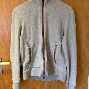 Grå kofta från Moncler - Säljer en äkta grå kofta från Moncler med ståkrage och hel dragkedja framtill. Koftan har två fickor med randiga dragkedjor och broderad Moncler-logga på ärmen. Ribbad mudd vid ärmslut och nederkant. Har en liten skavanka på vänster baksida, som är professionellt sydd av en skräddare 