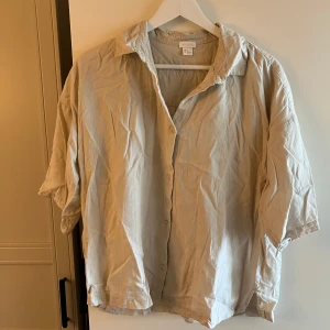 Beige linneskjorta från H&M - Säljer en oversized beige skjorta från H&M i svalt linnematerial. Skjortan har korta ärmar, klassisk krage och knäppning framtill. Perfekt för en chill och avslappnad stil, lätt att matcha med jeans eller shorts.