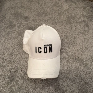 Vit Dsquared2 ICON keps - Snygg vit keps från Dsquared2 med svart broderad text 'ICON' framtill och 'DEAN & DAN CATEN' baktill. Justerbar rem med metallspänne för perfekt passform. Tillverkad i bomull, klassisk form och stilren design som sticker ut.