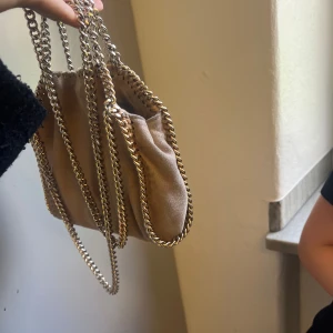 Beige axelväska Stella McCartney - Snygg beige axelväska från Stella McCartney med kedjedetaljer i silver runt kanterna och som axelrem. Väskan är i mjuk mocka-liknande syntet och har magnetlås samt innerfack med logotypmönster. Perfekt storlek för det viktigaste.