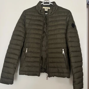 Olivgrön pufferjacka från Roco Baroco - Säljer en olivgrön pufferjacka från Roco Baroco. Jackan har quiltad design, två dragkedjefickor fram och en hög krage. Märkets logga sitter på ärmen och jackan har snygga detaljer i silver. Perfekt för kyliga dagar!