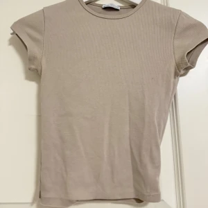 Beige ribbad t-shirt från Bershka - En stilren beige t-shirt från Bershka i ribbat material. Modellen är kortärmad med rund halsringning och har en slim passform som sitter snyggt på kroppen. Perfekt att matcha med jeans eller kjol för en clean look.