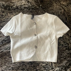 Vit croppad blus från H&M Divided - Säljer en vit croppad blus från H&M Divided med korta ärmar och tre snygga silvriga knappar framtill. Toppen har en lätt struktur i tyget och en enkel, stilren design som är perfekt för att lyfta din outfit.