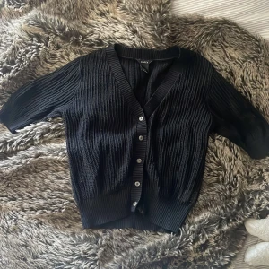 Svart ribbad kofta från Lindex - Svart kofta från Lindex i storlek S med ribbad stickning och snygga knappar framtill. Koftan har korta ärmar och v-ringad hals, perfekt att slänga på över en t-shirt eller linne. Materialet är stickat och ger en chill och stilren vibe.