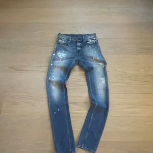 7 for all man kind jeans med slitningar och färgstänk - Säljer ett par blå jeans med coola slitningar och vita färgstänk. Materialet är jeans och de har en snygg tvättad effekt som ger en unik stil. Modell: Ronnie. Storlek: W29.                         Midja – 38 cm x 2 Höft – 45 cm x 2 Grenhöjd – 25 cm Ytterbenslängd/Innerbenslängd – 110/86 cm Lårvidd – 27 cm x 2 Benslut – 15 cm x 2 