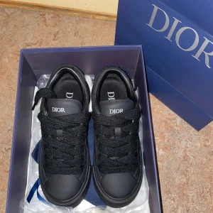 Svarta Dior sneakers i skinn - Snygga svarta Dior sneakers i skinn med diskret Dior-monogram präglat i lädret. Skorna har tjocka svarta snören, platt sula och rund tå. Klassisk låg modell med logotyp på plös och häl. Perfekta för dig som vill ha en lyxig och stilren look.