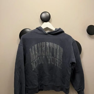 Mörkblå hoodie med Manhattan-tryck - Mörkblå hoodie med huva och glittrande text 'Manhattan New York' framtill i silvriga stenar. Tröjan har en avslappnad passform och långa ärmar. Perfekt för dig som gillar streetstyle och vill ha en snygg statementtröja.