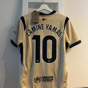 Barcelona Lamine Yamal tröja Nike S - Snygg Barcelona fotbollströja med Lamine Yamal och nummer 10 på ryggen. Tröjan är beige med svarta och blå detaljer, officiella loggor från Nike, Spotify och UNHCR samt klubbmärke på bröstet. Tillverkad i lätt och ventilerande polyester.