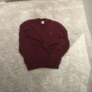 Vinröd stickad tröja från GANT - Vinröd stickad tröja från GANT med v-ringning och broderad logga på bröstet. Tröjan har långa ärmar och ribbade muddar vid ärmslut och nederkant. Perfekt för dig som gillar klassisk och stilren look.