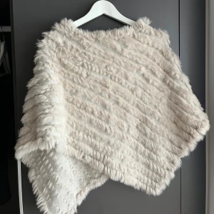 Beige pälsponcho från Natures Collection - Supermjuk och fluffig beige poncho från Natures Collection. Tillverkad i akryl och kaninpäls, med snygg struktur och asymmetrisk form. Perfekt för att hålla sig varm och se chic ut under kyliga dagar. Storlek S/M 
