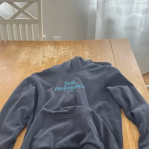 Mörkblå hoodie från Peak Performance - Mörkblå hoodie från Peak Performance med turkos broderad logga på bröstet. Klassisk känguruficka framtill och huva. Perfekt för dig som gillar en enkel och stilren look.