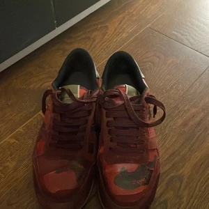 Valentino Rockrunner röda sneakers - Säljer ett par Valentino Rockrunner sneakers i rött och vinrött med coolt kamouflagemönster. Skorna har snörning, platt sula och detaljer i skinn och mocka. Snygga nitar baktill och svart hälparti ger extra edge. Perfekta för dig som vill sticka ut med lyxig streetstyle.