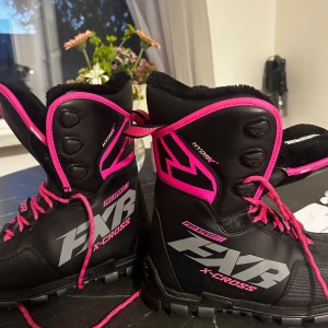 FXR X-Cross svarta och rosa boots - Säljer ett par svarta FXR X-Cross boots med starka rosa detaljer och snörning. Skorna har grov sula för bra grepp, högt skaft och är fodrade för extra värme. Perfekta för vintern och utomhusaktiviteter. Snygg design med tydliga loggor och kontrasterande färger. Passar 36 utan tjocka strumpor ny pris cirka 2000