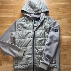 Beige jacket from Barbour - Säljer en beige jagka från Barbour International med huva och quiltad front. Jackan har mjuka ärmar, ribbade muddar och dragkedja framtill. Perfekt för dig som vill ha en stilren och bekväm jacka med snygg logga på ärmen.