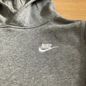 Grå hoodie från Nike med broderad logga - Klassisk grå hoodie från Nike med broderad vit logga på bröstet. Tröjan har huva, lång ärm och en stor magficka framtill. Tillverkad i mjuk bomullsblandning som är skön att ha på sig. Perfekt för chill dagar eller när du vill ha en enkel streetstyle.