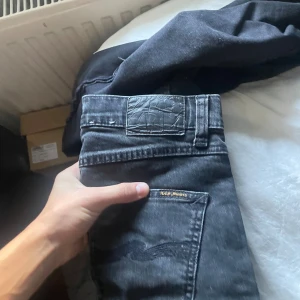 Svarta jeans från Nudie Jeans - Säljer ett par svarta jeans från Nudie Jeans med klassisk bakficka och broderad vågdetalj. Jeansen har en mörk tvätt och är tillverkade i mjukt denimtyg. Passar perfekt för dig som gillar stilrena och tidlösa jeans. W29 L32