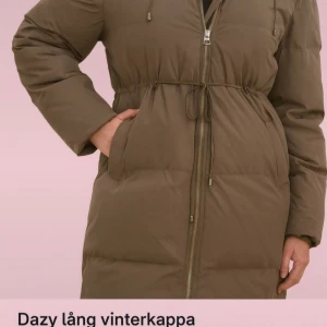 Olivgrön lång vinterkappa från Dazy - Lång, olivgrön vinterkappa från Dazy med quiltad design och dragsko i midjan för snygg passform. Jackan har huva, tvåvägsdragkedja och stora fickor framtill. Perfekt för kalla dagar när du vill hålla stilen och värmen.