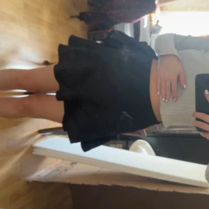 Svart omlottkjol med rosett - Supersöt svart kort omlottkjol med volang och stor rosett i sidan. Kjolen har en feminin och flowy passform som ger en snygg siluett. Perfekt att styla med crop top eller stickad tröja. Dragkedja i sidan för enkel påklädning.