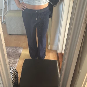 Mörkblå mjukisbyxor från Juicy Couture - Mörkblå mjukisbyxor från Juicy Couture med broderad logga på benet, snörning i midjan med guldfärgade detaljer och fickor med tryckknappar bak. Byxorna har en rak passform och är tillverkade i mjuk sammet för en lyxig känsla.