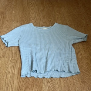 Ljusblå ribbad topp med volangkant - Säljer en ljusblå ribbad topp med korta ärmar och volangkant både vid ärmslut och nederkant. Toppen har en rund halsringning och är croppad för en trendig look. Perfekt att matcha med jeans eller kjol.