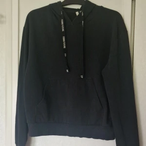 Svart hoodie med huva och känguruficka - Svart hoodie med texten Femme future tryckt på snörena och stor känguruficka framtill. Ribbad nederkant som har ett veck på mitten och den är litte tvättpåvrrkad i färgen (se bild). Perfekt för chill dagar och har en klassisk, enkel look med huva och snörning.