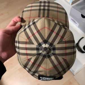 Burberry keps  - Snygg keps från Burberry med det ikoniska rutiga mönstret i beige, svart, vitt och röda linjer. Justerbar baktill med svart plastspänne och tydlig Burberry London England-patch. Tillverkad i bomull, perfekt för dig som gillar street och designer.