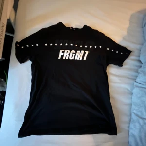  Moncler t-shirt - Svart t-shirt från Moncler x Fragment med vit FRGMT-print på bröstet och stjärnor längs bröstet och ärmarna. Klassisk passform och rund hals. Perfekt för dig som vill sticka ut med en snygg logga.