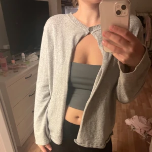 Ljusgrå kofta från Design by Si - Supermjuk ljusgrå kofta från Design by Si med enkel knapp högst upp vid halsen. Koftan har lång ärm, rak passform och är tillverkad i ett tunt, stickat material som känns skönt mot huden. Perfekt att slänga över en topp för en chill look.