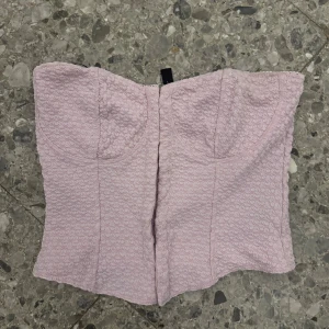 Rosa korsett från H&M Divided - Supersöt ljuslila korsett från H&M Divided med strukturerat tyg och hakknäppning framtill. Toppen är axelbandslös och har markerade kupor för en snygg siluett. Perfekt att styla med jeans eller kjol för en trendig look.