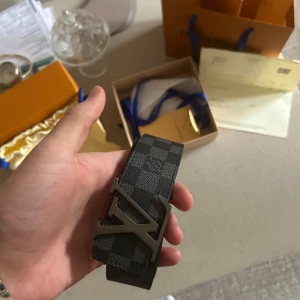 Louis Vuitton bälte - Snyggt Louis Vuitton bälte i klassiska Damier Graphite-mönstret. Bältet är svart och grått med rutigt mönster och har en stor LV-logga i metall som spänne. Kommer med originalpåse och box. Storlek: 90/36. Lätt använd och i bra skick