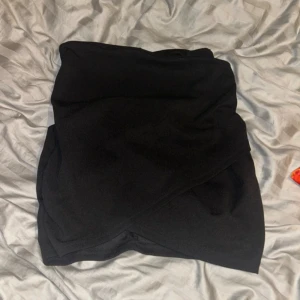 Svart omlottkjol i mjukt tyg - Säljer en svart kort omlottkjol i mjukt tyg. Kjolen har en enkel och stilren design med omlott-detalj framtill som ger en snygg look. Perfekt att matcha med en crop top eller stickad tröja. Passformen är normal och kjolen är helsvart utan mönster.